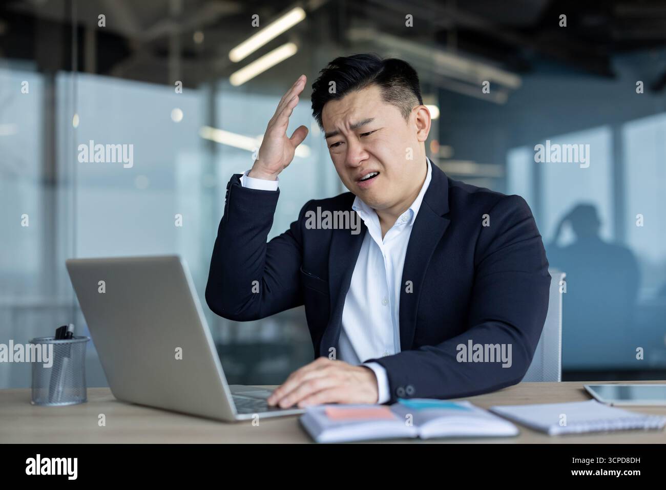 Frustré et contrarié jeune homme asiatique assis dans le bureau au bureau et travaillant sur ordinateur portable faisant des gestes avec la main. Banque D'Images
