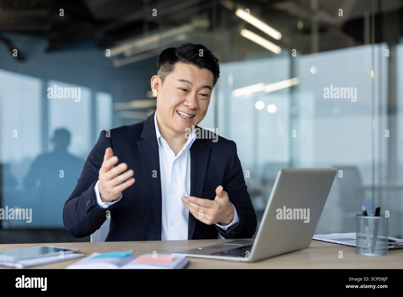 Jeune homme d'affaires asiatique assis dans le bureau au bureau dans un costume et parlant sur un appel vidéo sur un ordinateur portable, faisant des gestes avec ses mains. Banque D'Images