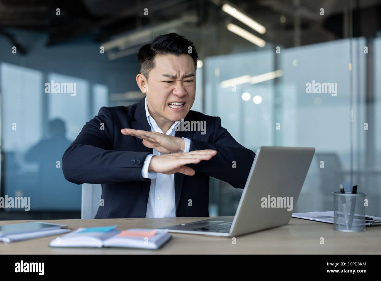 Jeune homme d'affaires asiatique en colère, employé de bureau assis dans le bureau devant l'ordinateur portable, parlant sur un appel vidéo et montrant émotionnellement le signe de déni avec les mains. Banque D'Images