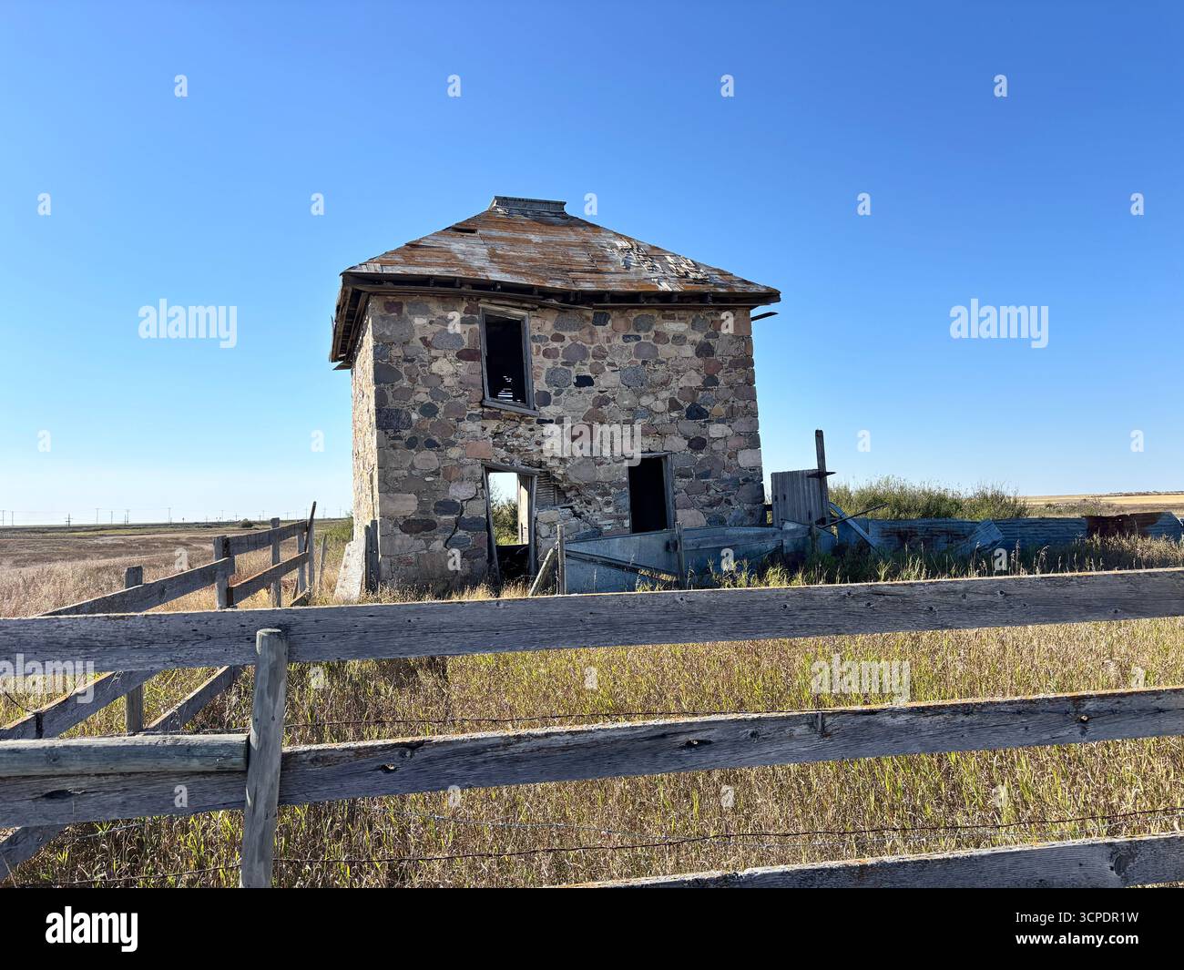 Vue d'une ferme en pierre abandonnée avec une clôture en bois altérée et de l'herbe des prairies envahie par la végétation sous un vaste ciel bleu dans la Saskatchewan rurale, Canada. - Image de stock capturée avec un smartphone