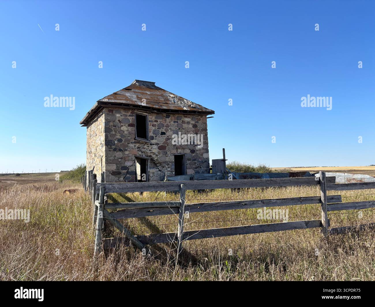 Vue d'une ferme en pierre abandonnée avec une clôture en bois altérée et de l'herbe des prairies envahie par la végétation sous un vaste ciel bleu dans la Saskatchewan rurale, Canada. - Image de stock capturée avec un smartphone