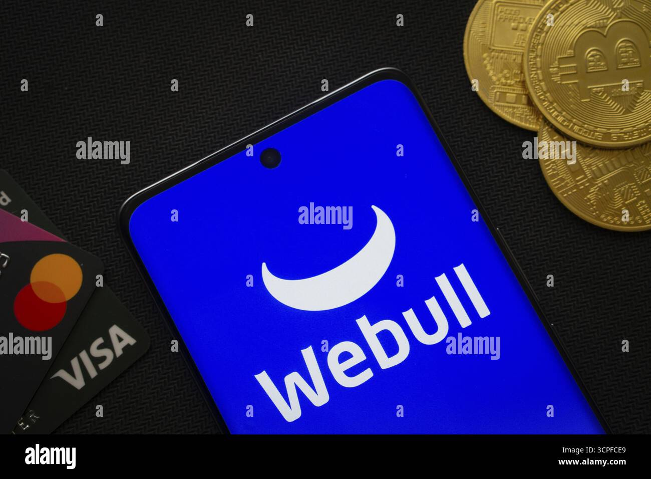 Dhaka, Bangladesh- 26 août 2025 : smartphone affichant le logo webull entouré de pièces Bitcoin et de cartes de paiement Banque D'Images