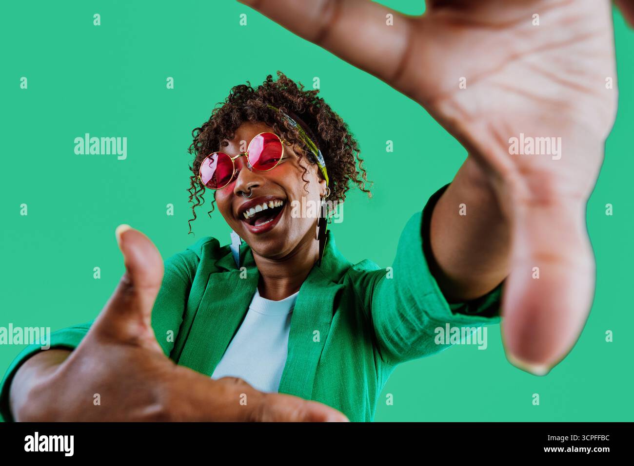 Jeune mannequin souriant tout en encadrant son visage avec les mains, portant une veste verte élégante et des lunettes de soleil roses ludiques, profitant d'un moment amusant agains Banque D'Images