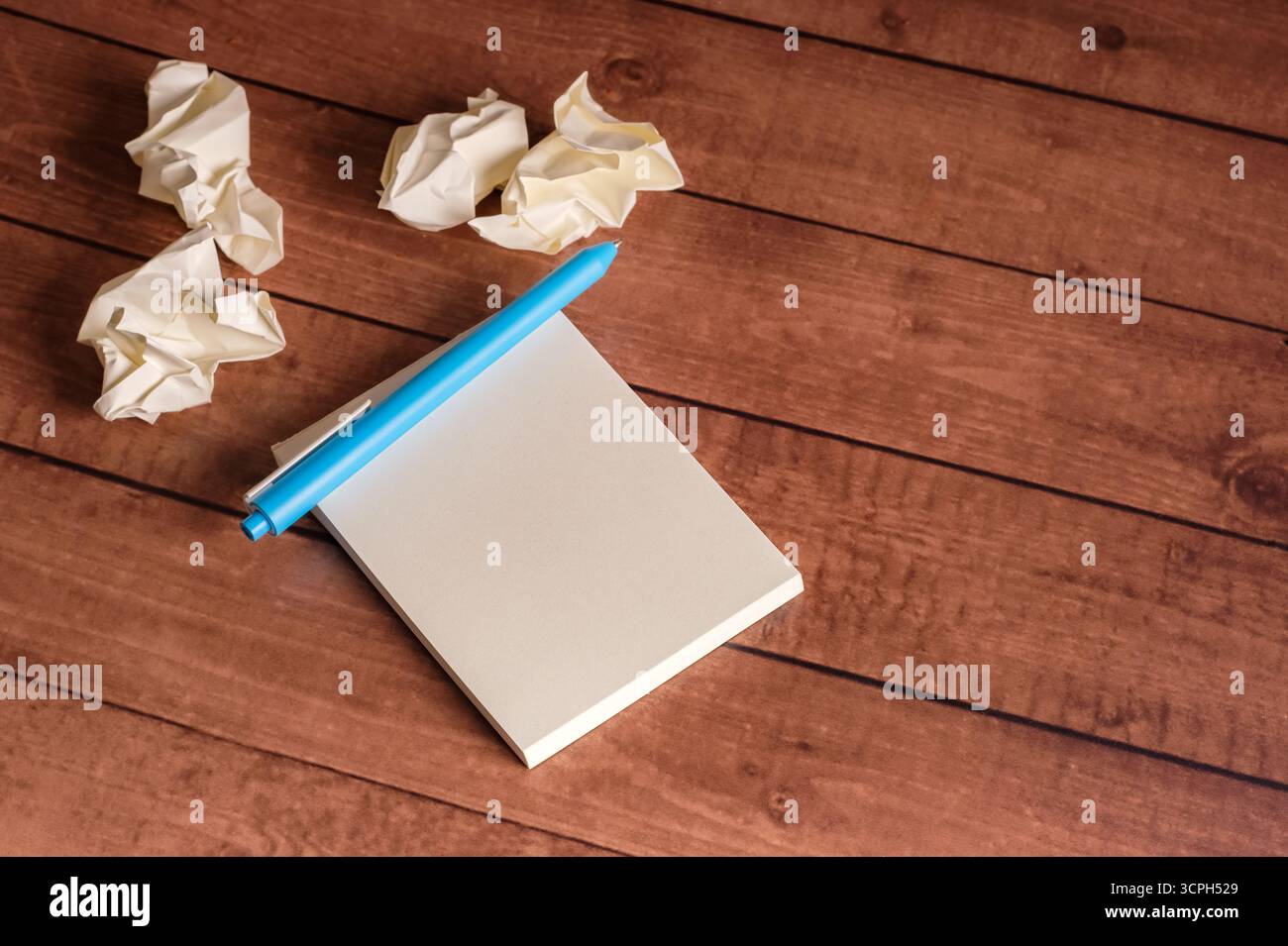 Bloc-notes vierge avec stylo bleu sur une table en bois entourée de boules de papier froissées. Espace de travail créatif pour le brainstorming et la génération d'idées. Banque D'Images