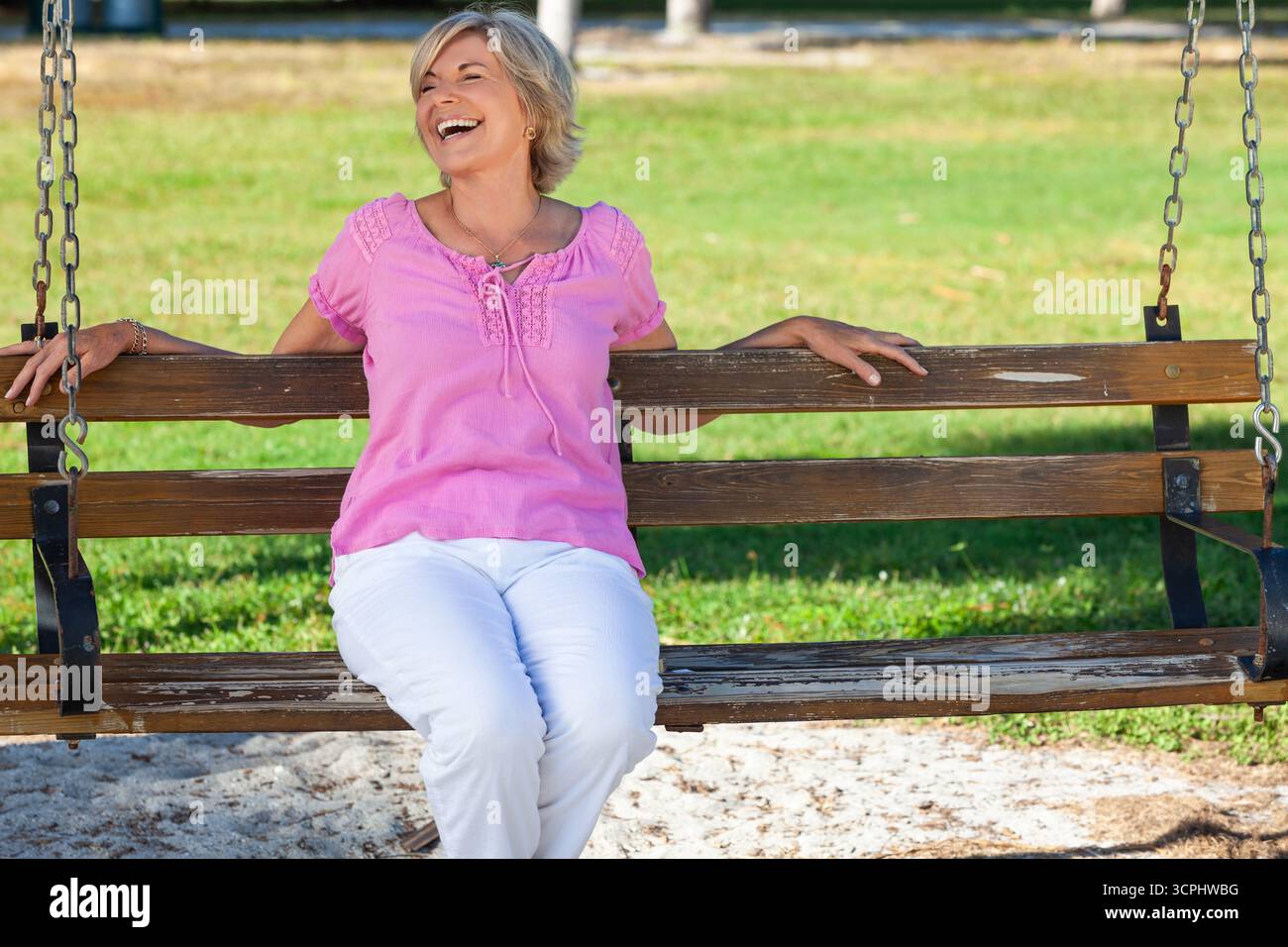 Portrait d'une belle femme âgée heureuse et en bonne santé assise à l'extérieur sur un banc riant avec de l'herbe verte derrière elle. Banque D'Images