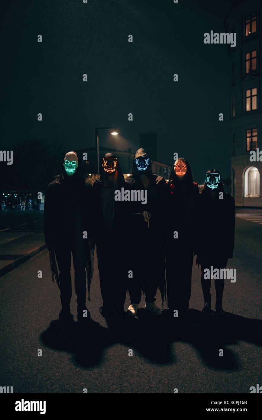 Un groupe de personnes portant des masques lumineux brillants se tient dans une rue sombre de la ville la nuit, avec des lumières et des ombres colorées pour un mystère étrange Banque D'Images