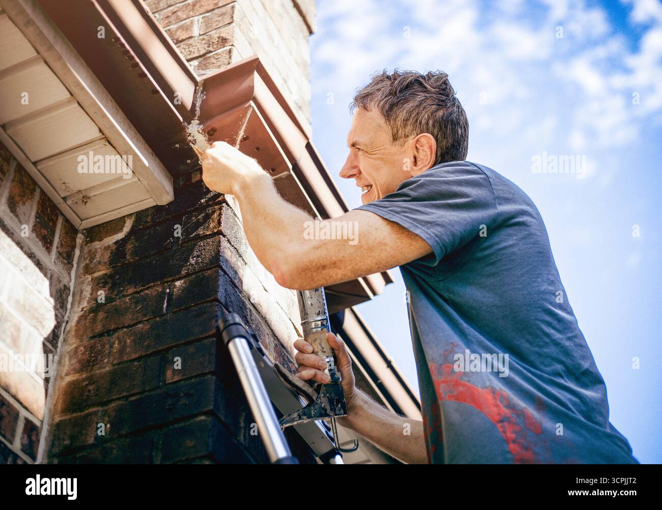 Un homme scelle une gouttière sur le toit d'une maison en briques surplombant un ciel bleu. Banque D'Images