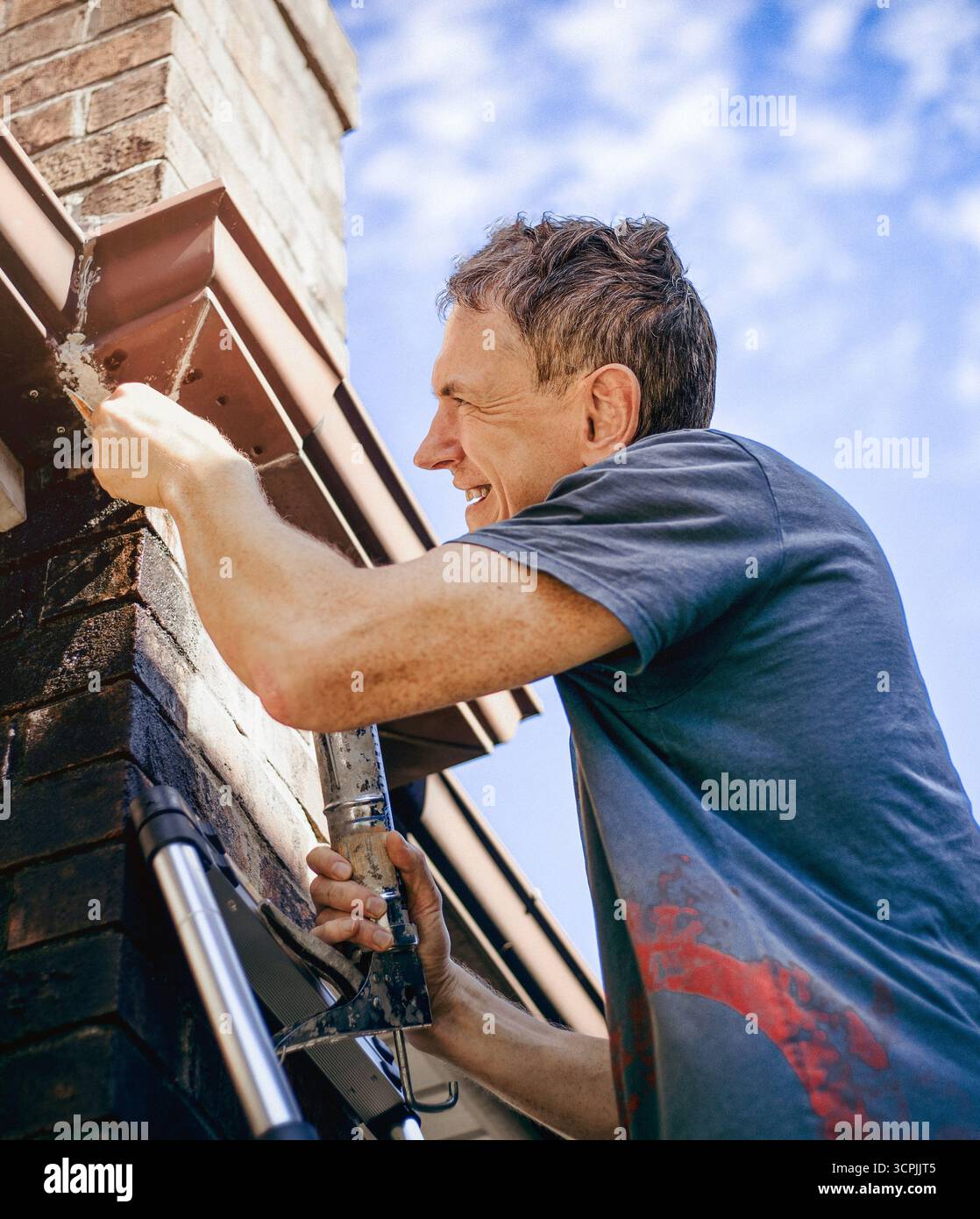 Un homme scelle une gouttière sur le toit d'une maison en briques surplombant un ciel bleu. Banque D'Images