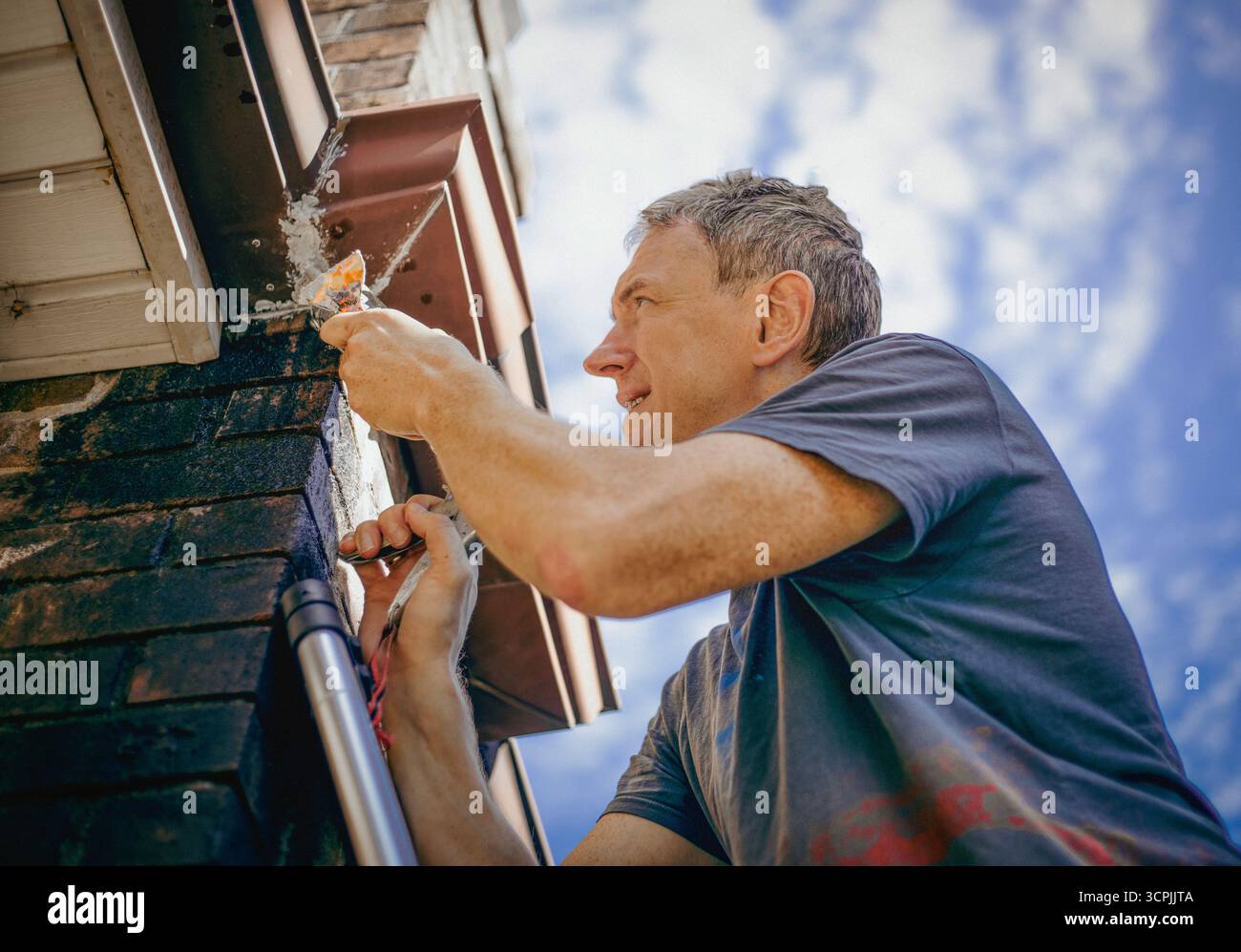 Un homme scelle une gouttière sur le toit d'une maison en briques surplombant un ciel bleu. Banque D'Images