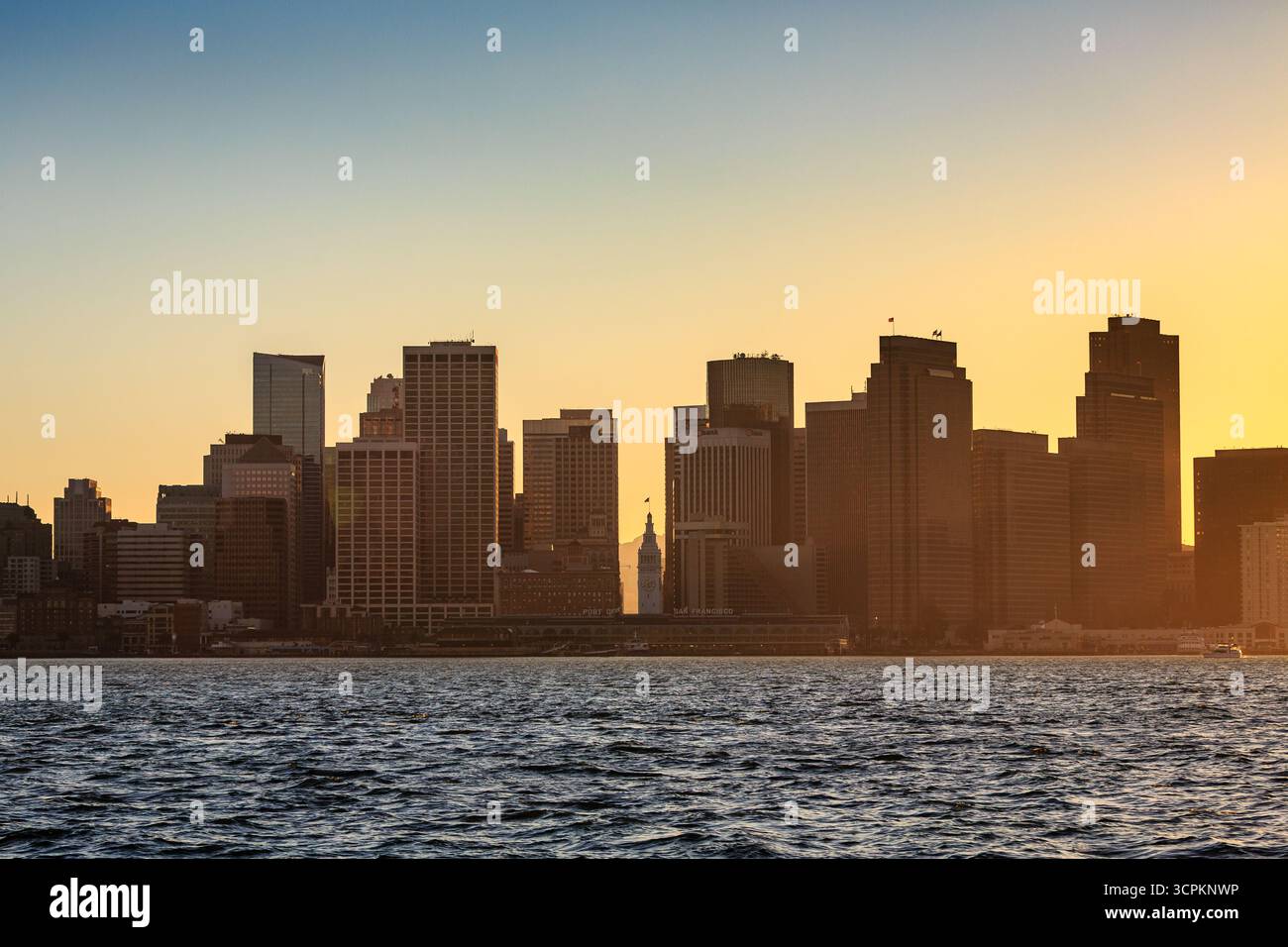 Une ligne d'horizon de San Francisco se silhouette contre un ciel dégradé au coucher du soleil, avec l'eau de la baie ondulant au premier plan. Banque D'Images