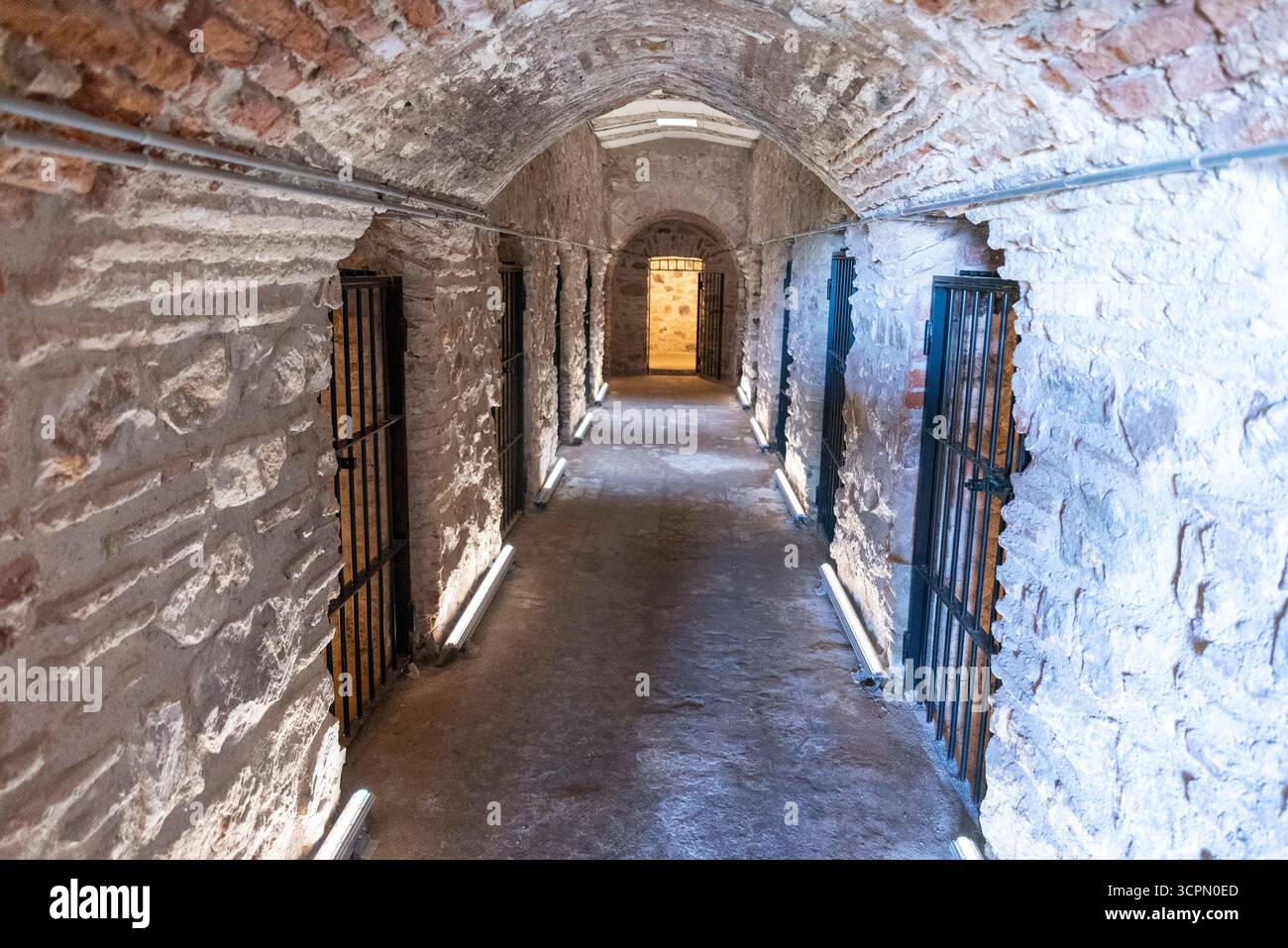 Un couloir étroit présente des murs en pierre brute et des portes barrées, mettant en valeur un cadre carcéral historique. L'éclairage moderne met en valeur les contours, creati Banque D'Images