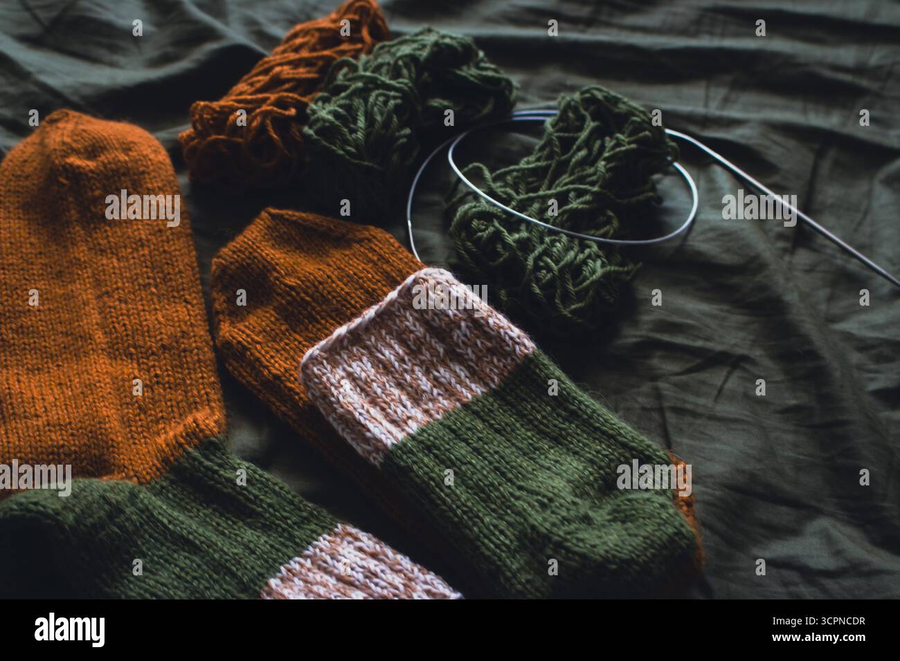 Chaussettes tricotées confortables avec boule de fil et aiguilles à tricoter. Chaussettes rayées en laine sur drap vert foncé. Couleurs d'automne douillettes. Crochet tissé gros plan. Banque D'Images