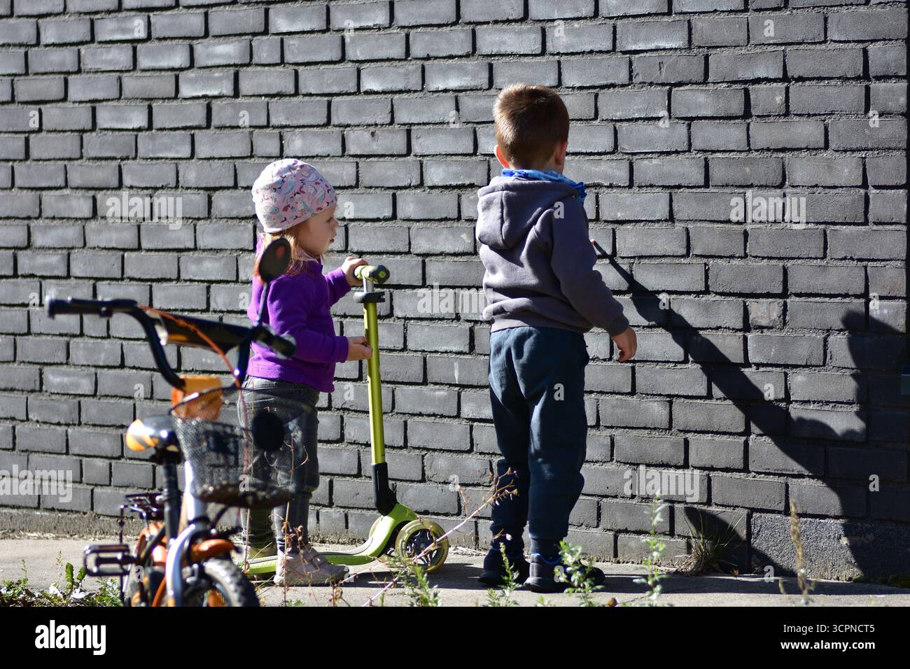 Garçon et fille avec scooter debout près du mur de briques grises, regardant les ombres. Curiosité infantile, découverte et exploration urbaine en plein air. Frères et sœurs p Banque D'Images