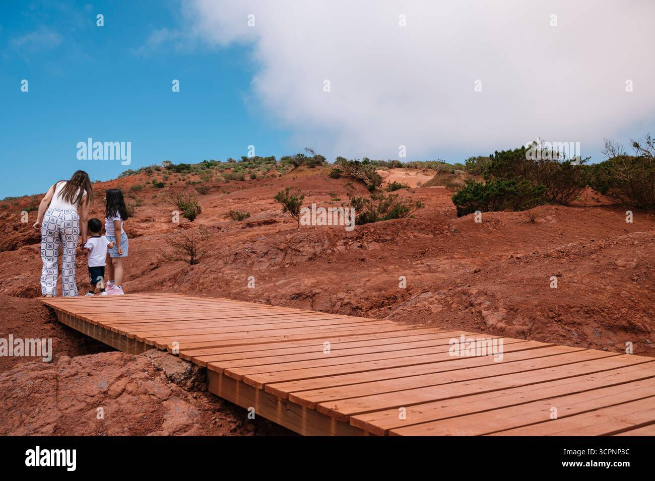 Une femme et deux enfants marchent sur un pont en bois sur un chemin de terre rouge. Le ciel est clair et le soleil brille Banque D'Images
