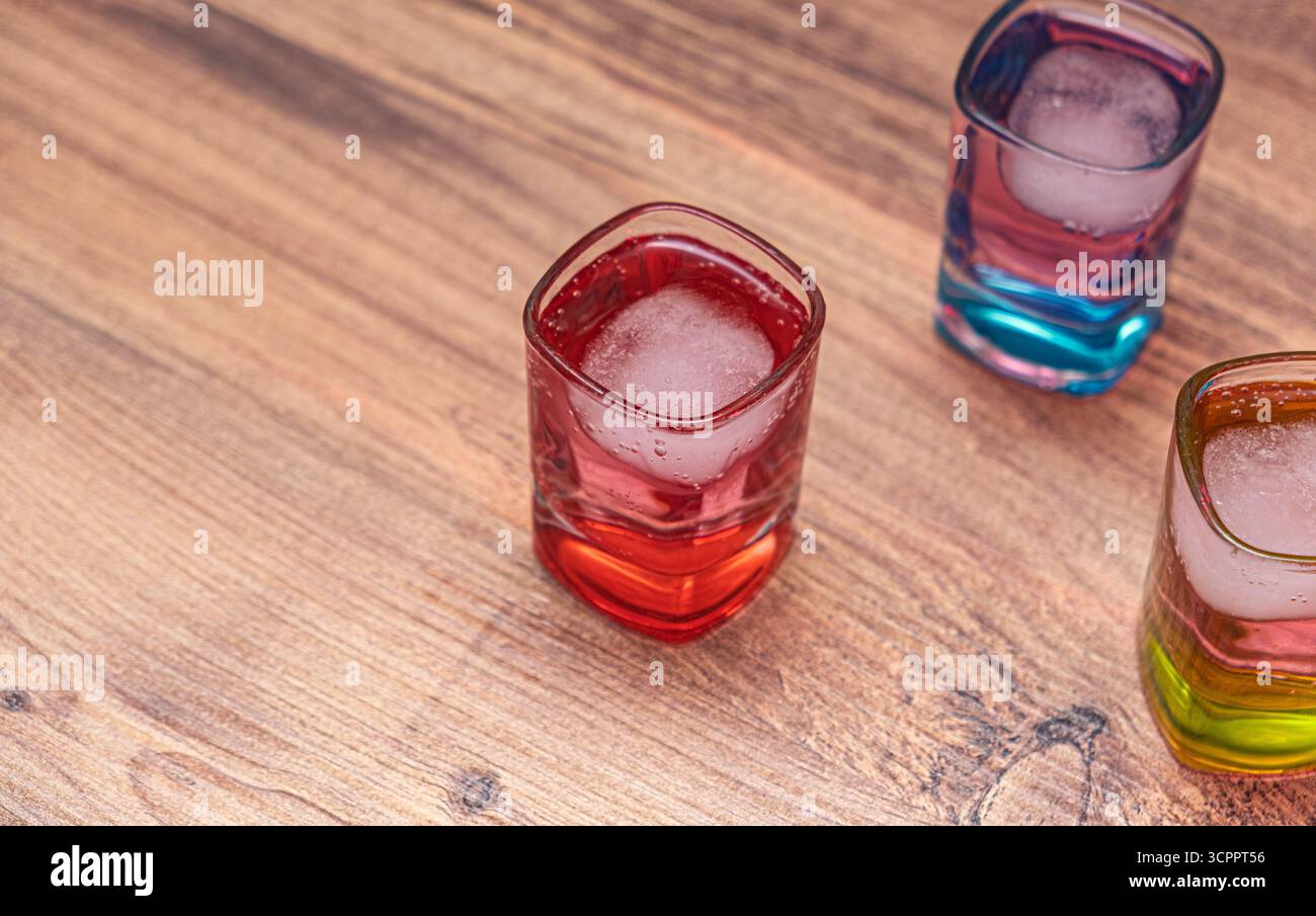 Trois verres de forme carrée remplis de boissons colorées et de glaçons, placés sur une surface en bois rustique. L'accent est mis principalement sur la boisson rouge. Banque D'Images