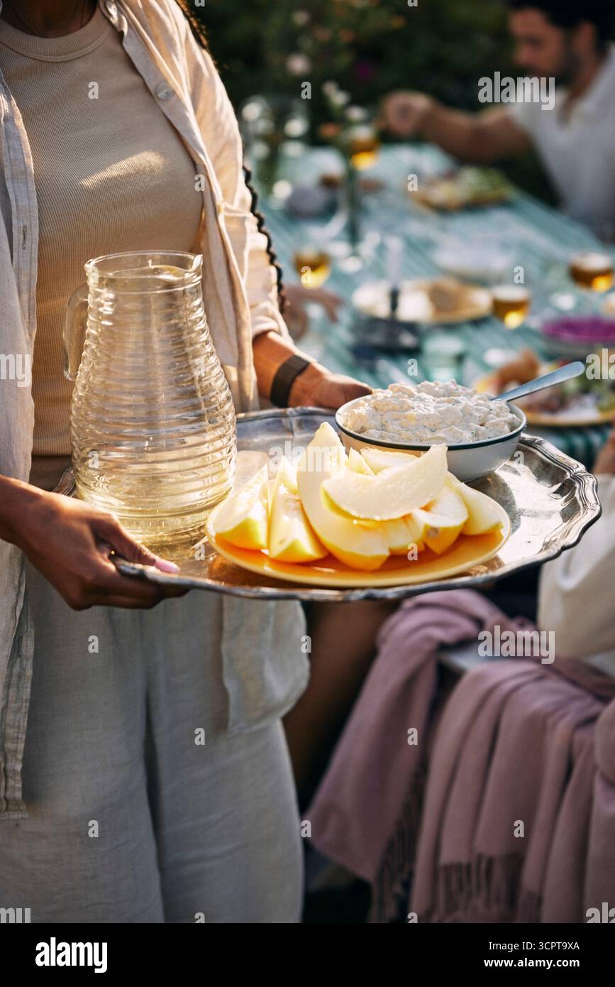 Section médiane de la femme tenant un plateau de melon avec pichet et saladier au dîner Banque D'Images