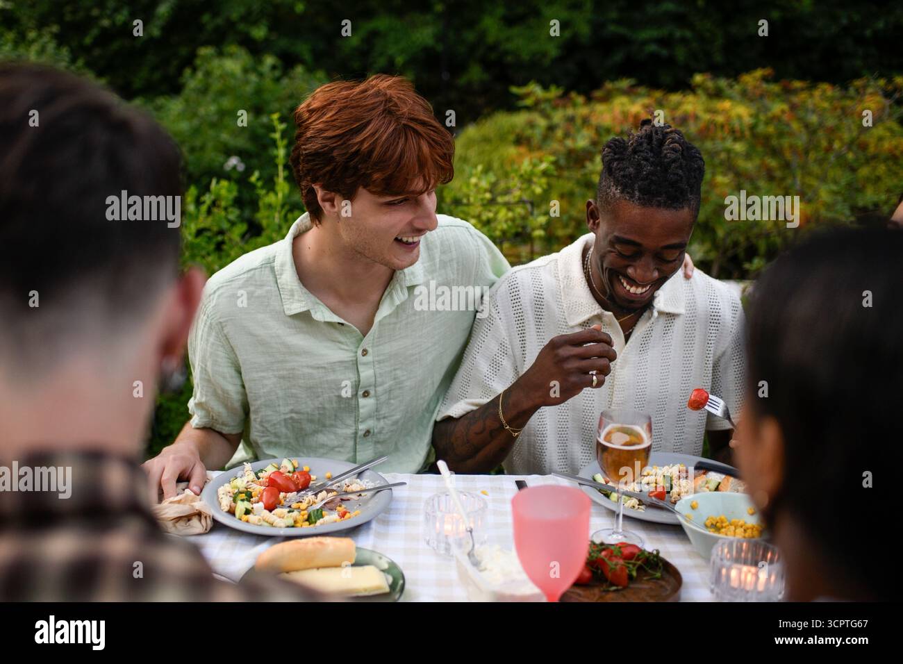 Jeune homme souriant assis avec le bras autour d'un ami mâle mangeant de la salade de pâtes au dîner dans la cour arrière Banque D'Images
