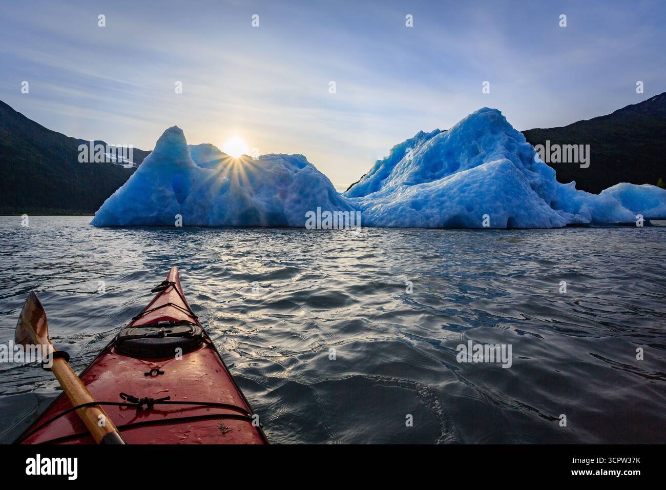 Un kayakiste pagaie vers un grand iceberg dans un plan d'eau, avec le soleil brillant derrière lui, créant une scène naturelle étonnante. Banque D'Images