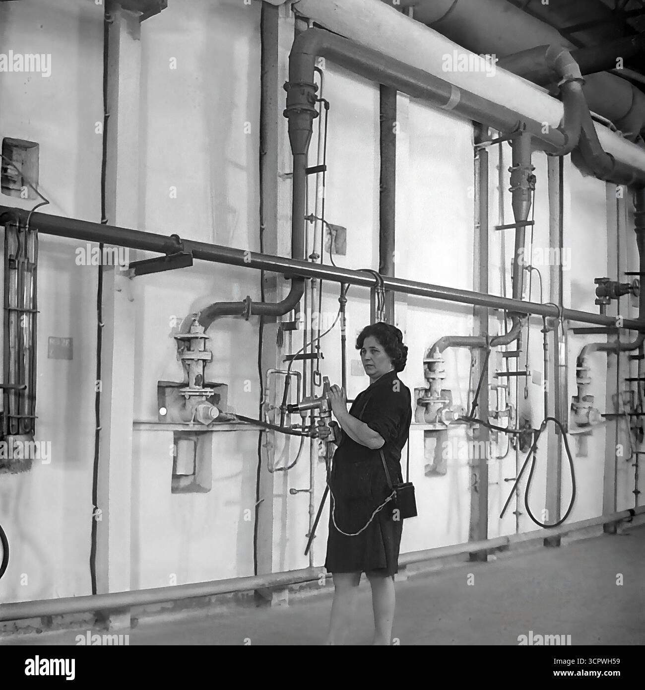 Une ingénieure utilise un pyromètre optique portable pour mesurer la température à l'intérieur d'un four industriel à l'usine de raccords et d'isolateurs de Slavyansk (AIZ) dans la RSS d'Ukraine dans les années 1970 Cette photo d'archives monochrome capture un moment crucial du processus de contrôle qualité dans la fabrication des isolants céramiques. La scène, placée contre un mur complexe de tuyaux et de jauges, met en évidence l'expertise technique et le rôle vital de spécialistes qualifiés dans l'industrie lourde de la région du Donbass, un fournisseur clé pour le réseau d'électrification de l'URSS Banque D'Images