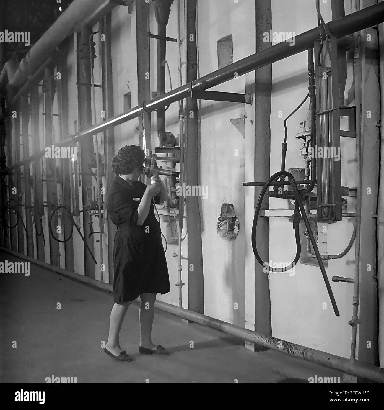 Une ingénieure utilise un pyromètre optique portable pour mesurer la température à l'intérieur d'un four industriel à l'usine de raccords et d'isolateurs de Slavyansk (AIZ) dans la RSS d'Ukraine dans les années 1970 Cette photo d'archives monochrome capture un moment crucial du processus de contrôle qualité dans la fabrication des isolants céramiques. La scène, placée contre un mur complexe de tuyaux et de jauges, met en évidence l'expertise technique et le rôle vital de spécialistes qualifiés dans l'industrie lourde de la région du Donbass, un fournisseur clé pour le réseau d'électrification de l'URSS Banque D'Images