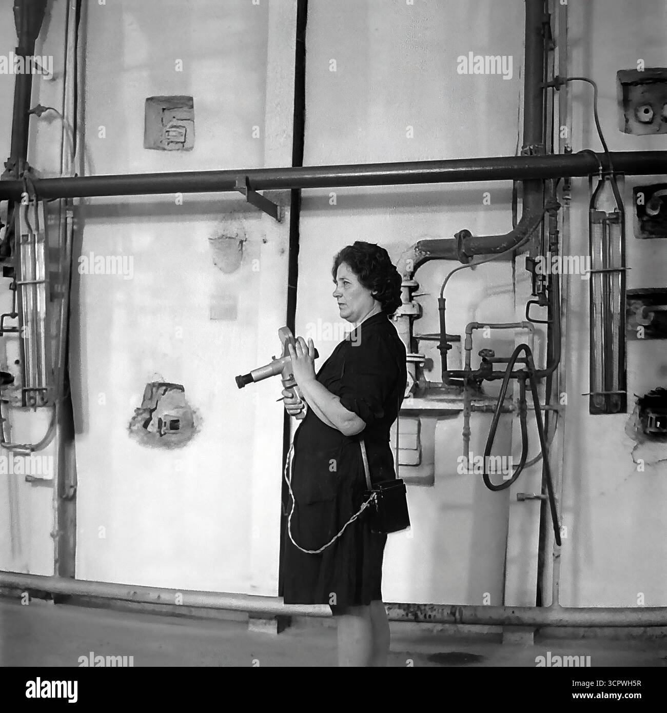 Une ingénieure utilise un pyromètre optique portable pour mesurer la température à l'intérieur d'un four industriel à l'usine de raccords et d'isolateurs de Slavyansk (AIZ) dans la RSS d'Ukraine dans les années 1970 Cette photo d'archives monochrome capture un moment crucial du processus de contrôle qualité dans la fabrication des isolants céramiques. La scène, placée contre un mur complexe de tuyaux et de jauges, met en évidence l'expertise technique et le rôle vital de spécialistes qualifiés dans l'industrie lourde de la région du Donbass, un fournisseur clé pour le réseau d'électrification de l'URSS Banque D'Images