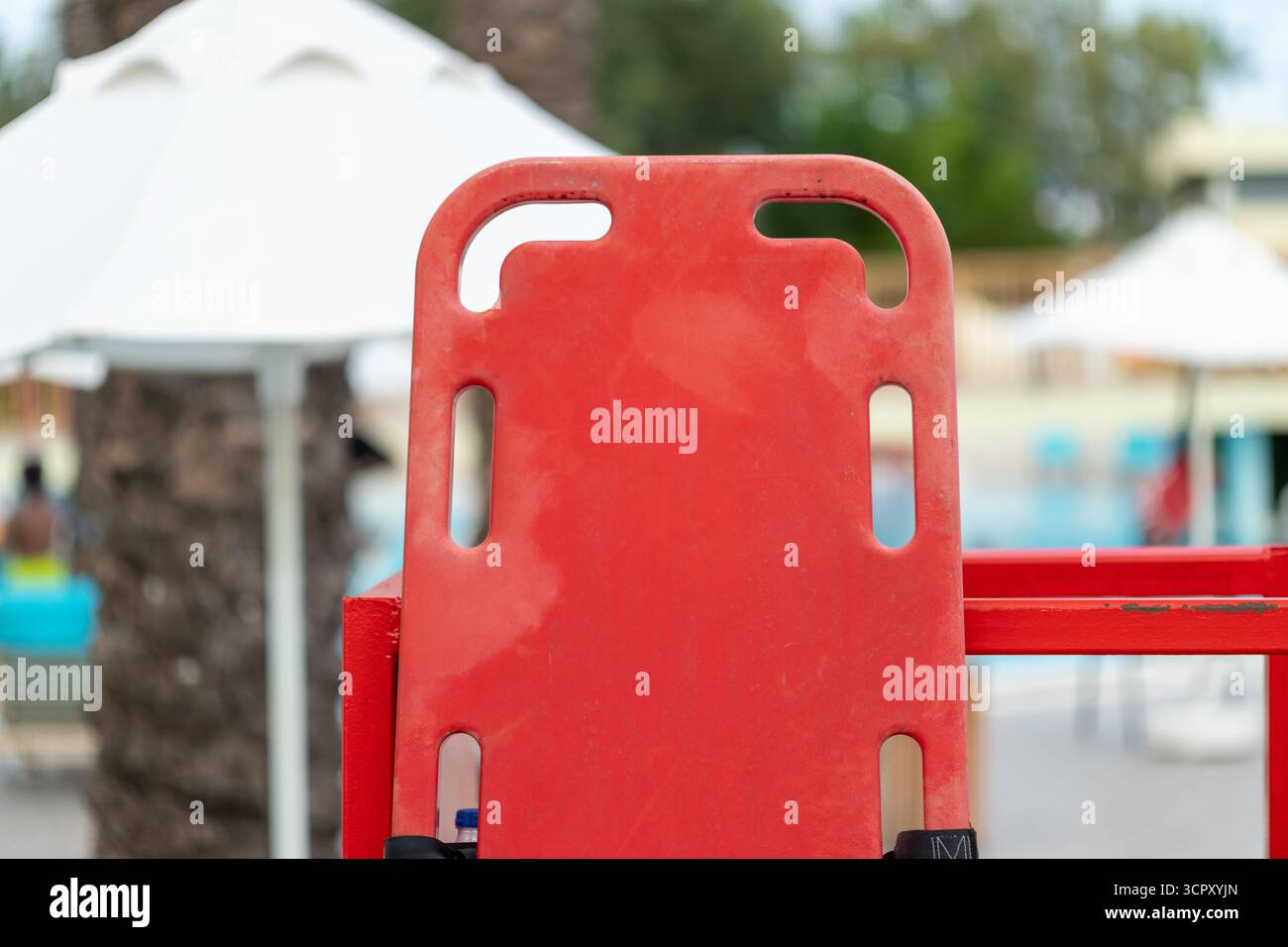 Gros plan d'une planche dorsale rouge avec des découpes de poignée, montée sur un support rouge. L'arrière-plan présente des parasols flous et une piscine. Banque D'Images