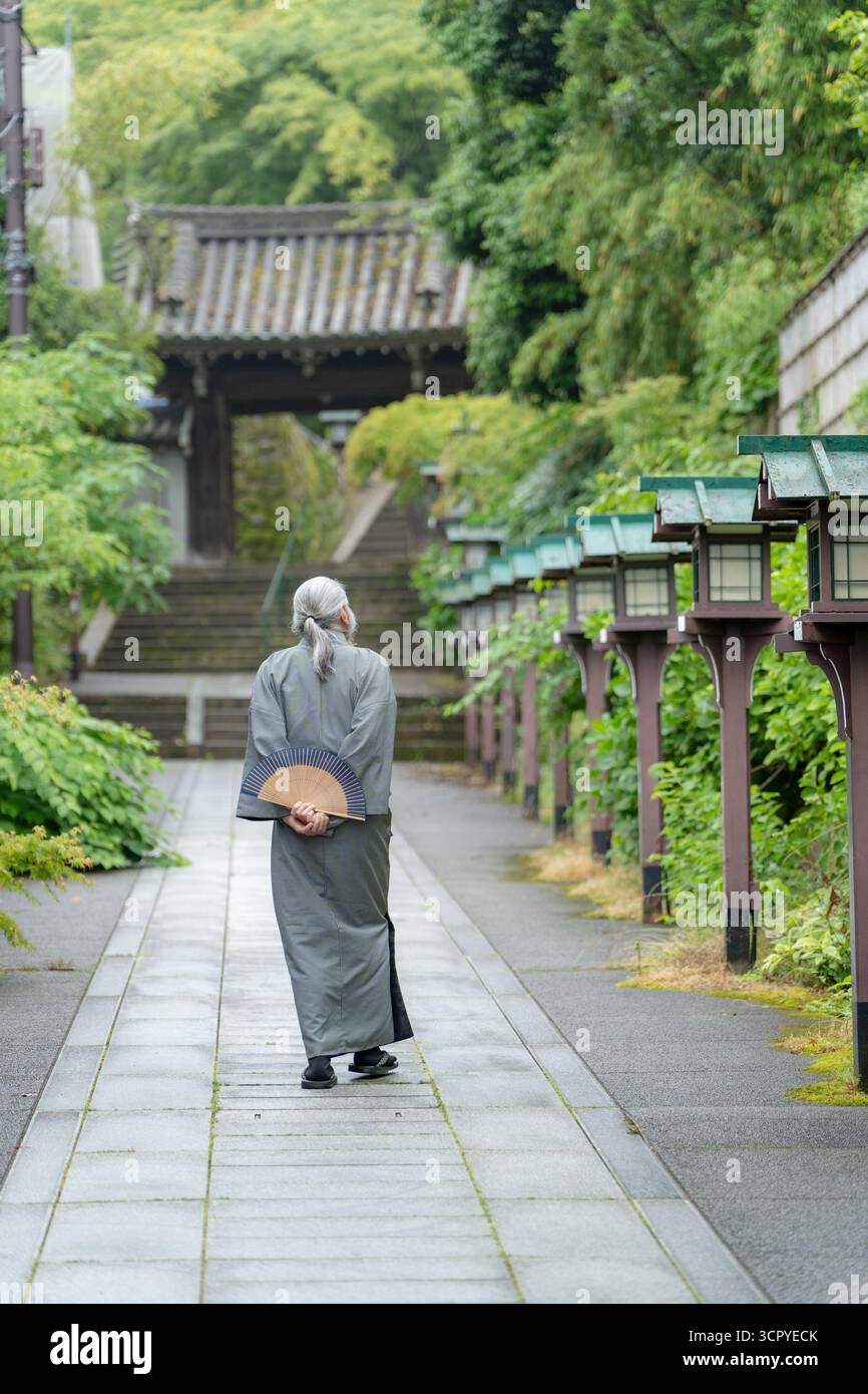 Un vieil homme dans un kimono tenant un ventilateur pliant marche sur un chemin de pierre vers une porte de temple. Kyoto, Japon. Banque D'Images