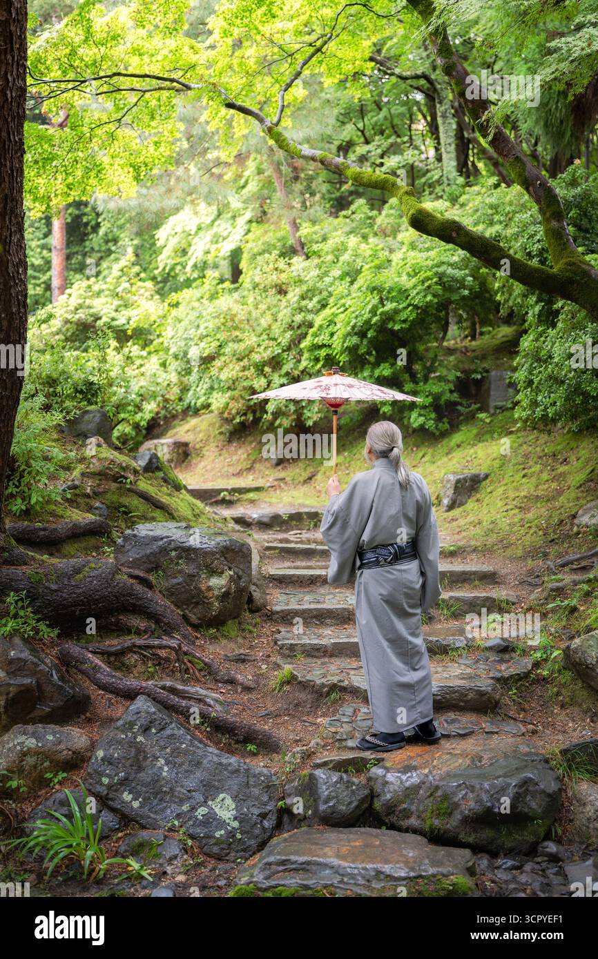 Un homme âgé dans un kimono tenant un parapluie japonais traditionnel monte les marches en pierre moussue dans une forêt. Kyoto, Japon. Banque D'Images