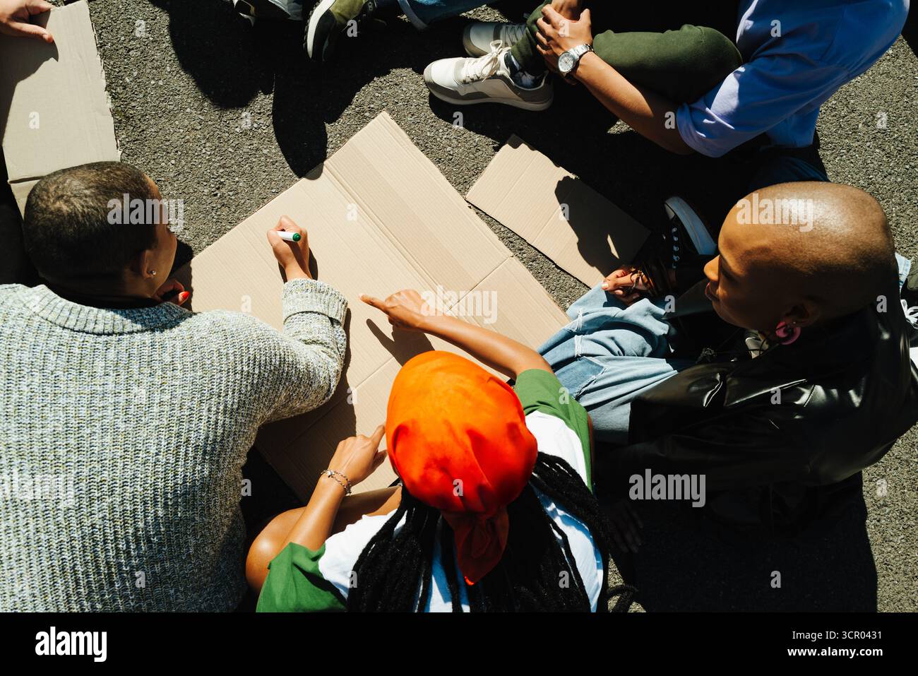 Un groupe diversifié de personnes s'assoit ensemble, créant des panneaux de protestation sur le carton à la lumière du jour. La scène capture leur effort collaboratif et leur concentration, sym Banque D'Images