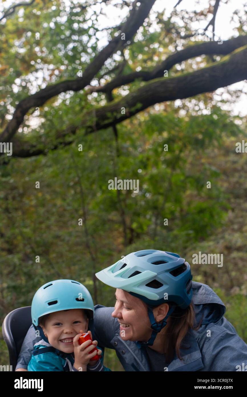Mère souriante et enfant dans des casques bleus profitant d'une pause collation pendant la promenade à vélo dans la forêt d'automne avec un fond de bokeh naturel. Concept de cycli familial Banque D'Images