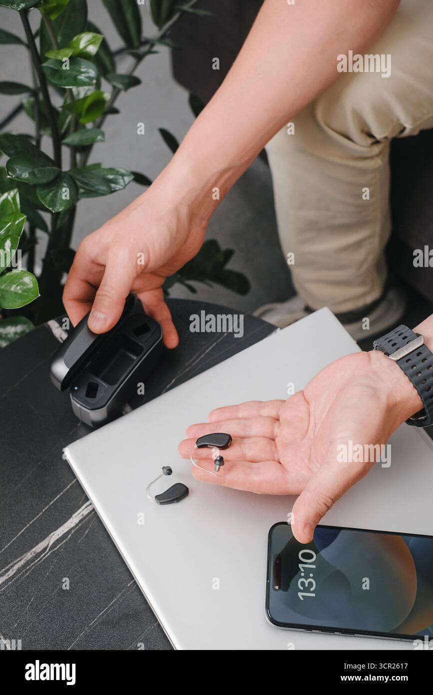 Aides auditives numériques modernes, smartphone et étui de chargement sur une table de bureau élégante, avec une personne préparant les appareils pour le travail. Environnement professionnel Banque D'Images