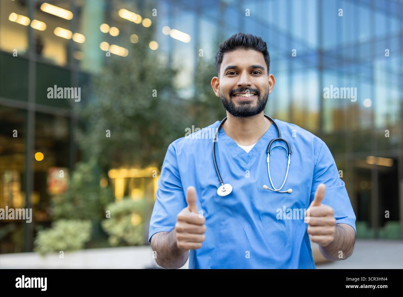 Jeune médecin indien souriant portant des gommages bleus et un stéthoscope, debout à l'extérieur devant un bâtiment moderne, montrant un signe pouce vers le haut positif avec les deux mains Banque D'Images