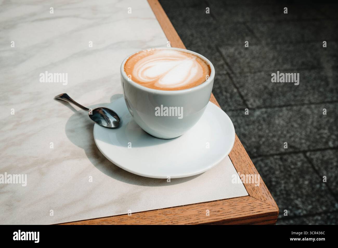 gros plan d'un macchiato servi dans une tasse blanche sur une soucoupe avec une cuillère, montrant un design de coeur dans la mousse, posé sur une table de café en plein air sous la lumière du jour Banque D'Images