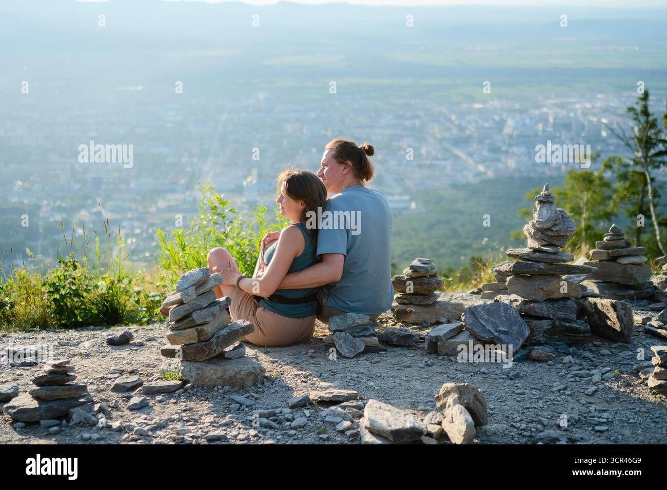 Homme et femme assis embrassés de derrière tout en profitant d'une vue urbaine spectaculaire pendant le voyage de randonnée, piles de pierre artistiques en face. Voyage et romance Banque D'Images