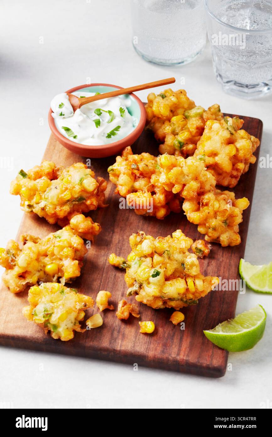 Beignets de maïs croustillants sur une planche de bois avec un côté de sauce crémeuse et des quartiers de citron vert. Banque D'Images