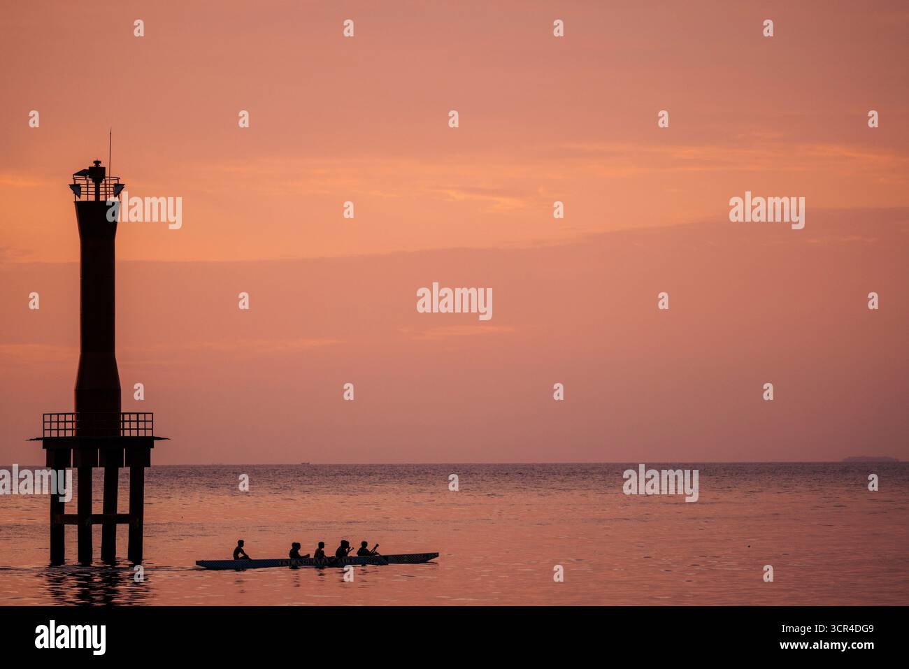 Des personnes silhouettées rament un canoë sur l'eau calme près d'un phare au coucher du soleil. Padang, Sumatra Ouest, Indonésie Banque D'Images