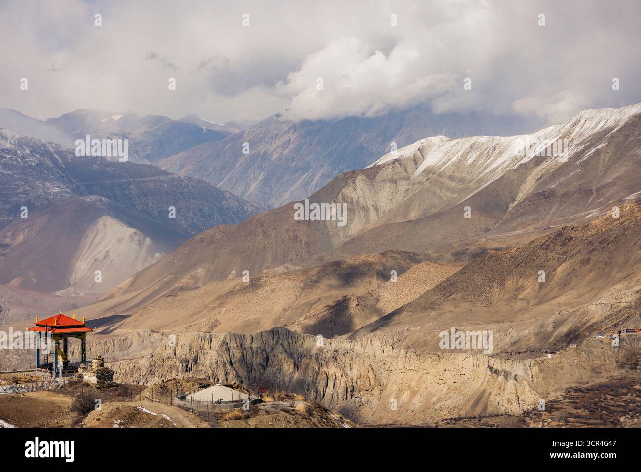Vaste paysage montagneux avec des nuages spectaculaires et une petite structure avec un toit rouge. Muktinath, Annapurna, district de Mustang, province de Gandaki, Népal Banque D'Images