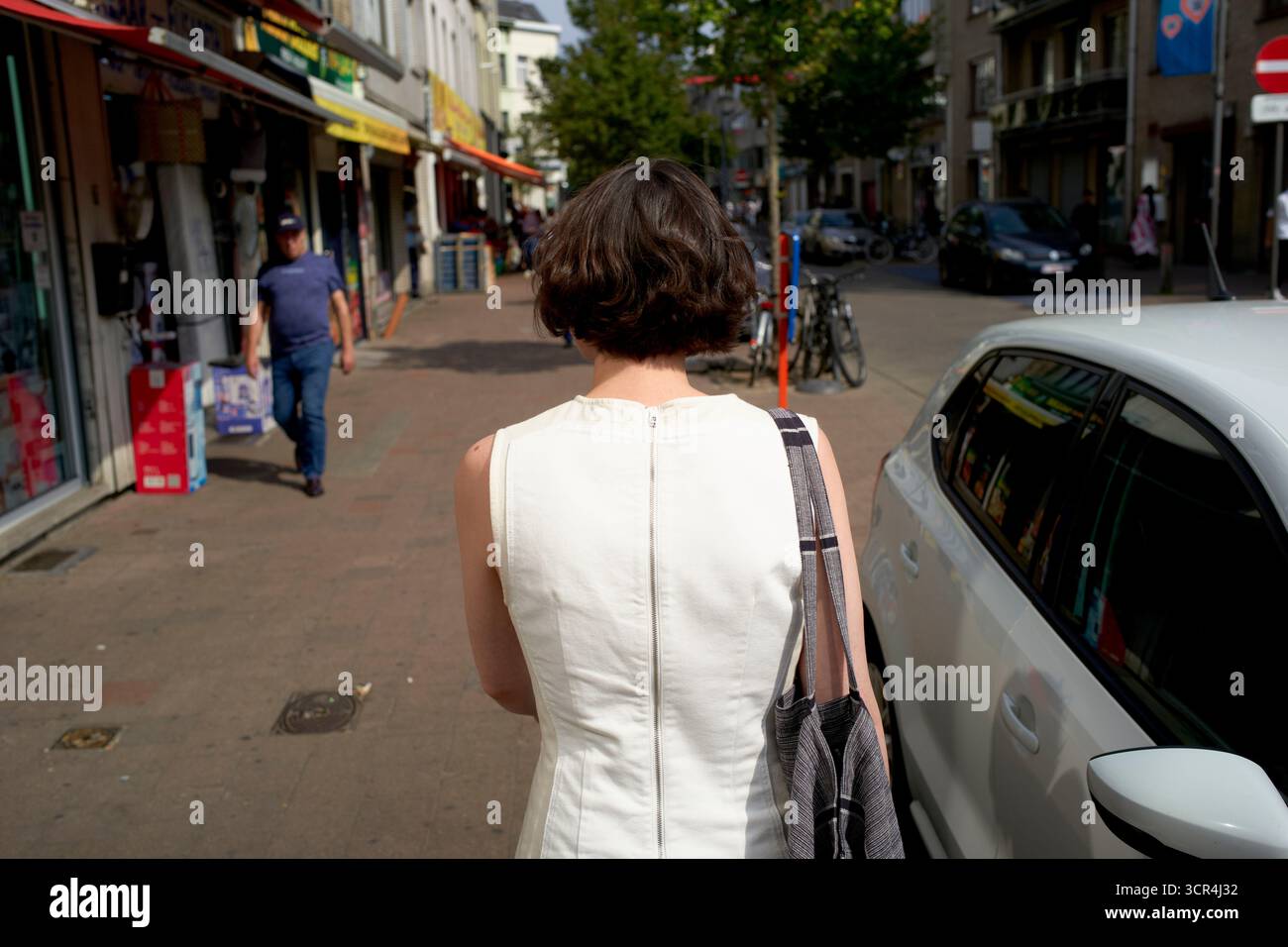 Femme en robe blanche marchant dans une rue animée de la ville avec des magasins et une voiture garée. Allemagne Banque D'Images