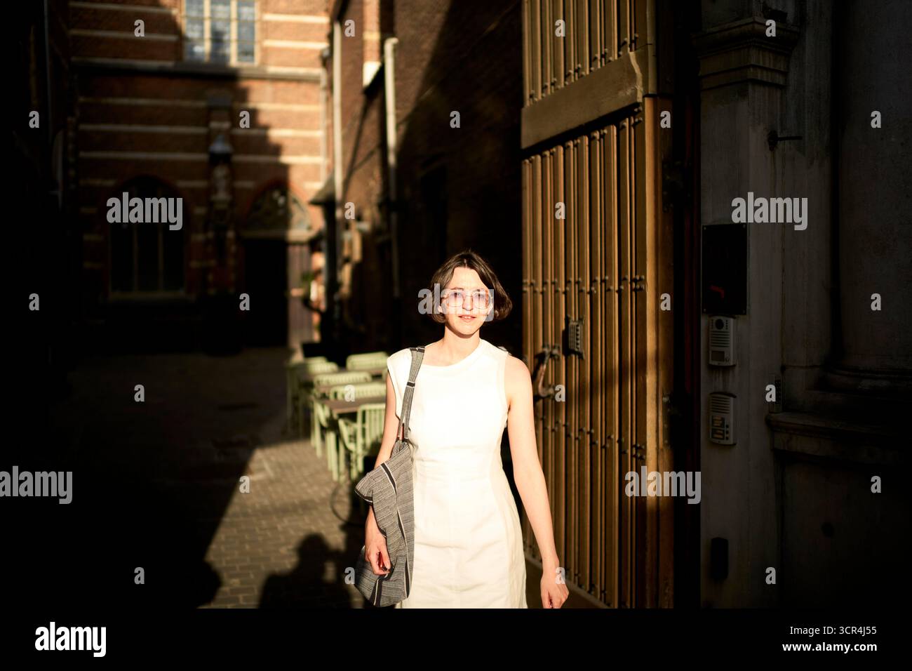 Jeune femme dans une robe blanche marchant dans une allée urbaine ensoleillée avec des bâtiments en briques. Allemagne Banque D'Images
