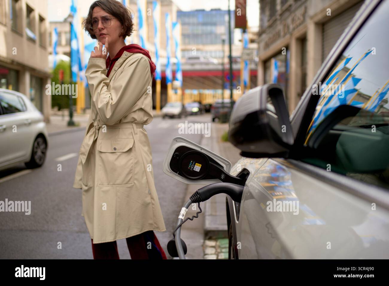Femme dans un trench-coat se tient près d'une voiture électrique se chargeant dans une rue de la ville. Bruxelles, Belgique Banque D'Images