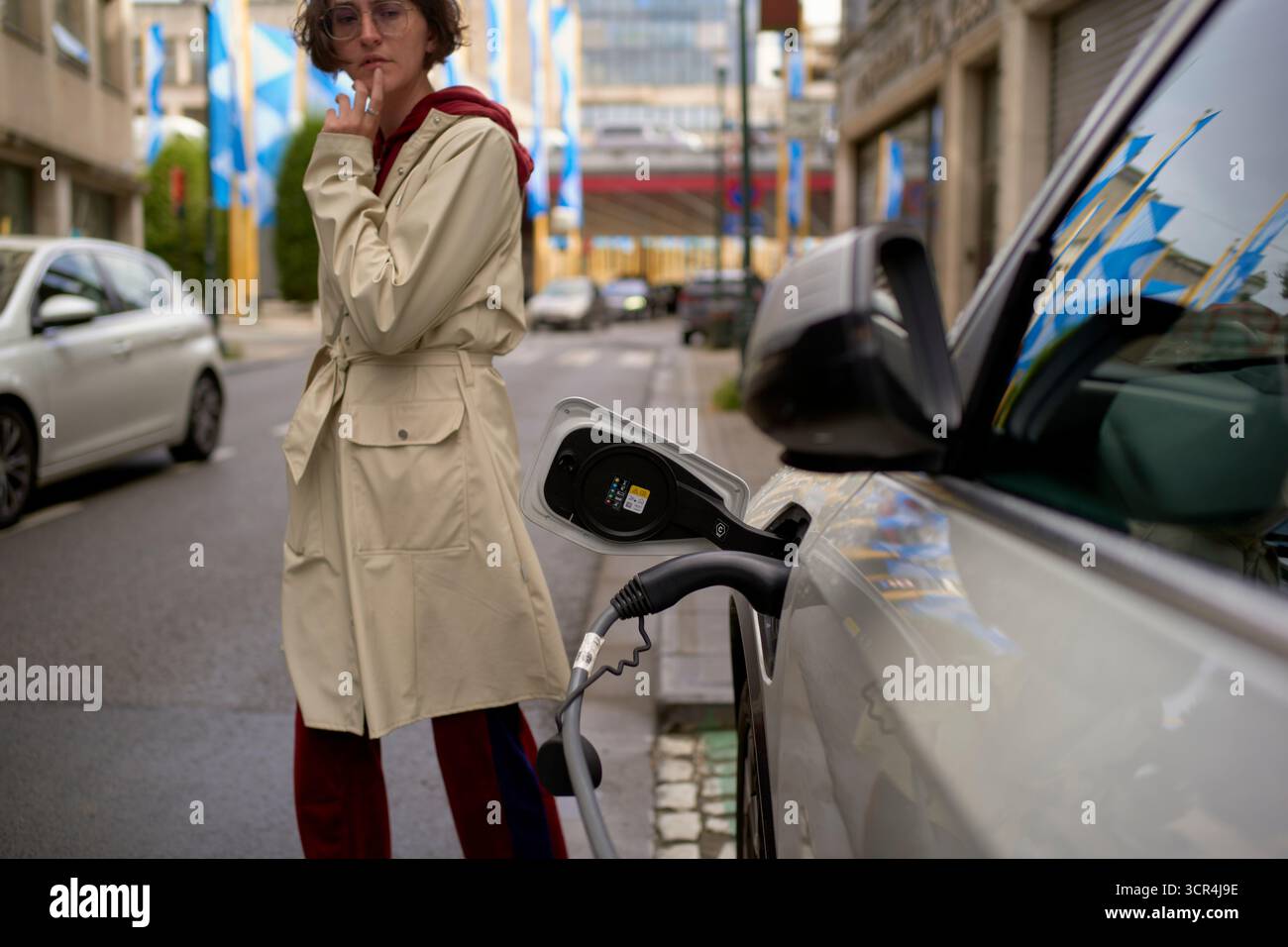 Femme dans un trench-coat se tient près d'une voiture électrique se chargeant dans une rue de la ville. Bruxelles, Belgique Banque D'Images