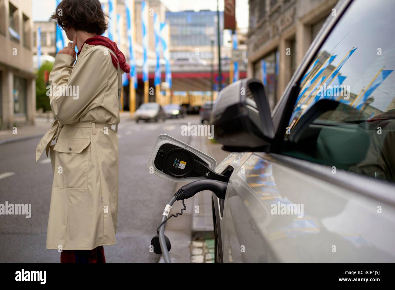 Personne en manteau beige debout près d'une voiture électrique se chargeant sur une rue urbaine. Bruxelles, Belgique Banque D'Images