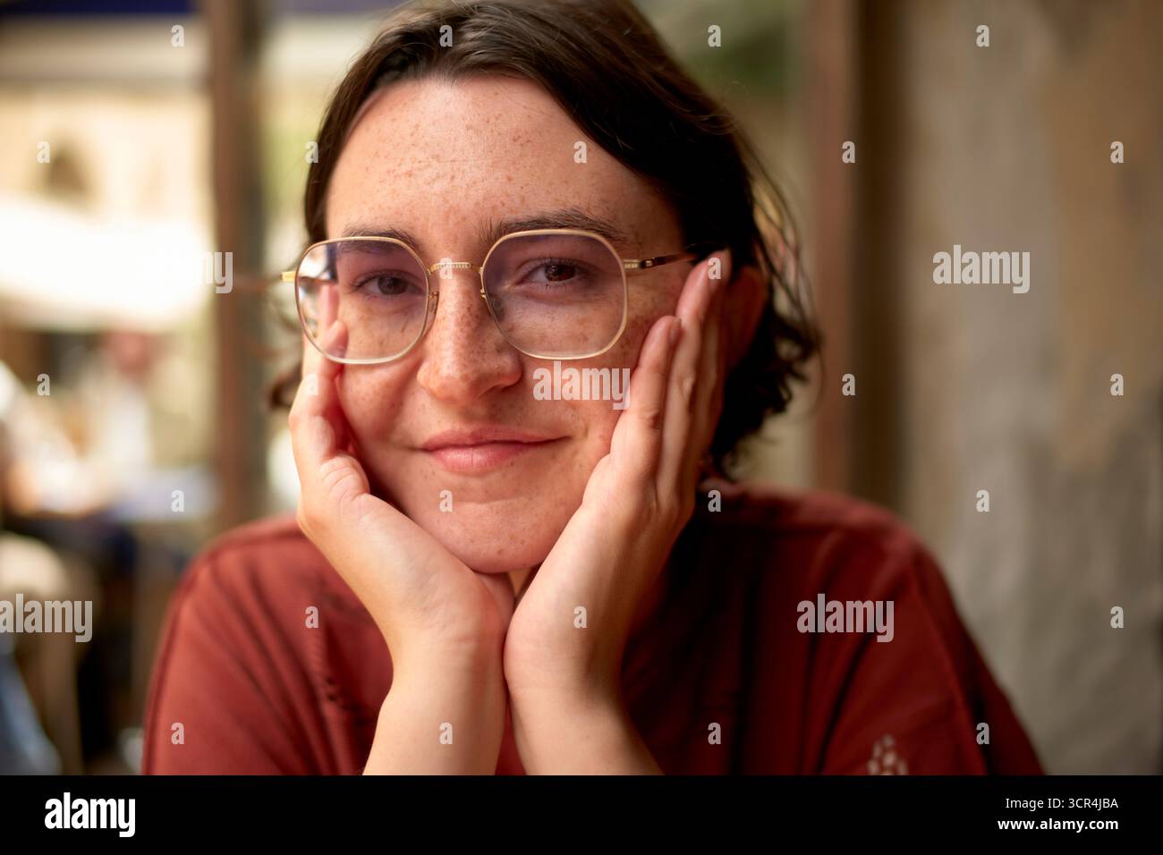 Jeune femme avec des lunettes et des cheveux courts souriants, les mains sur les joues, dans un cadre décontracté. Marseille, France Banque D'Images