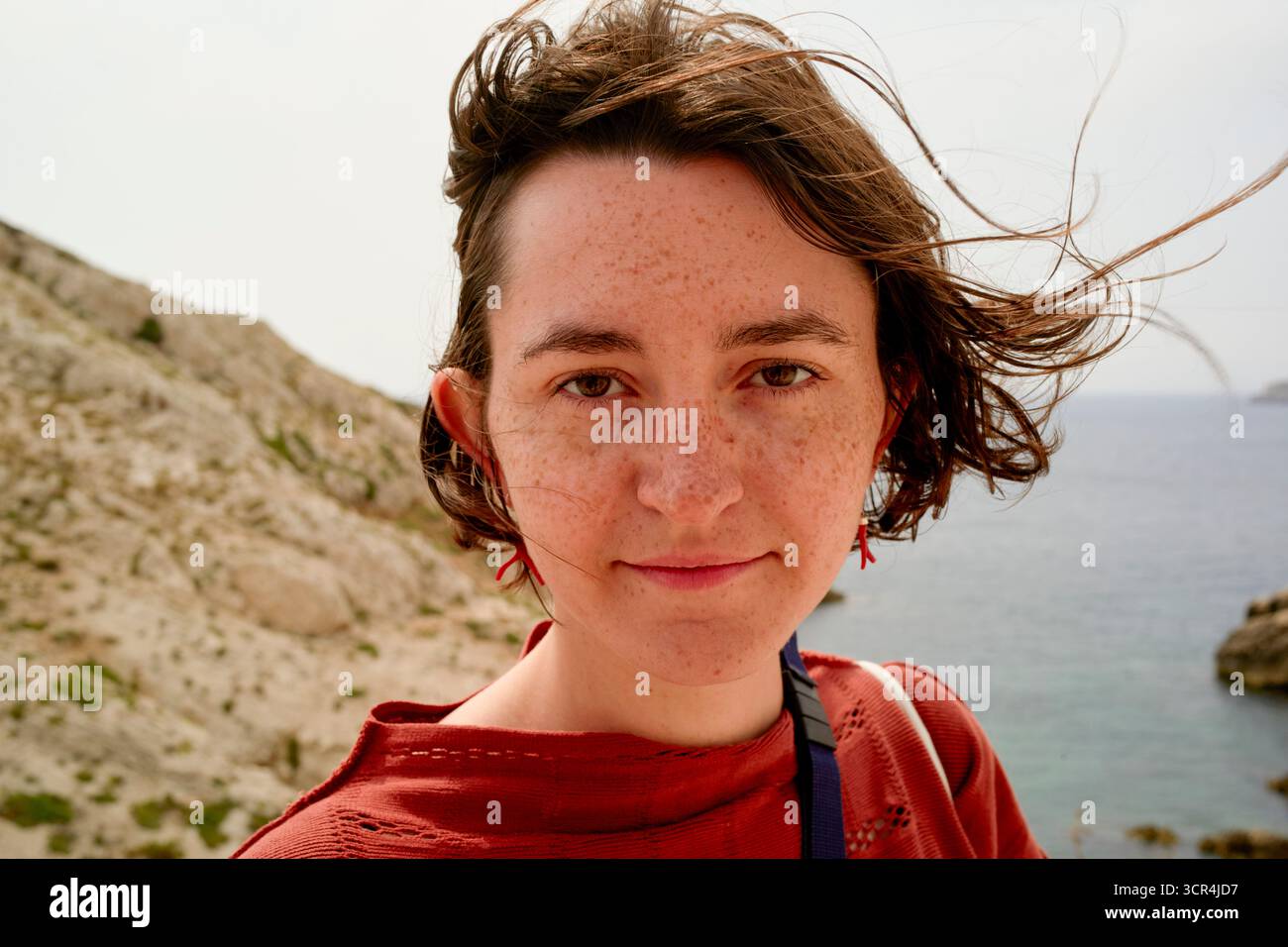 Femme en chemise rouge sourit avec les cheveux balayés par le vent par la mer et le paysage rocheux. Marseille, France Banque D'Images