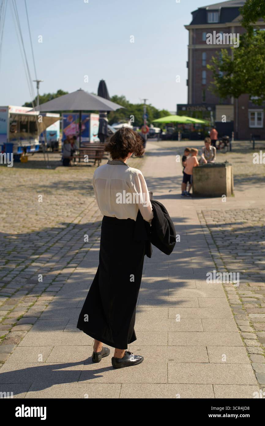 Femme aux cheveux courts marche à l'extérieur par une journée ensoleillée tenant une veste, regarde jouer aux enfants. Mer du Nord, Allemagne Banque D'Images
