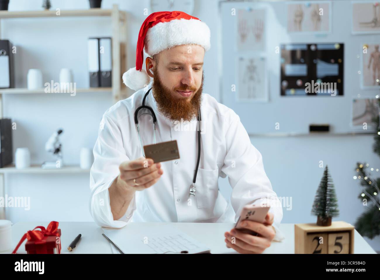 Docteur homme en uniforme et chapeau de Père Noël tenant la carte de crédit et utilisant un smartphone pendant le quart de travail la veille de Noël. copier l'espace Banque D'Images