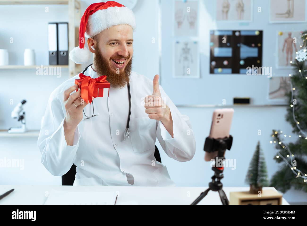 Docteur en chapeau de Père Noël tenant une boîte-cadeau rouge montrant les pouces vers le haut regardant sur l'appareil photo de smartphone. Étudiant en médecine travaillant dans le quart de travail à Noël ou nouvel an Banque D'Images