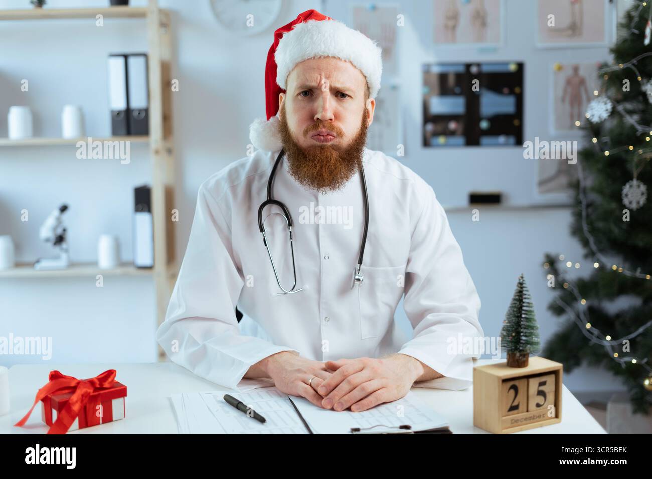 Portrait de docteur barbu dans le chapeau du Père Noël coulant les lèvres et les joues travaillant sur le quart de travail à l'hôpital la veille de Noël. copier l'espace Banque D'Images