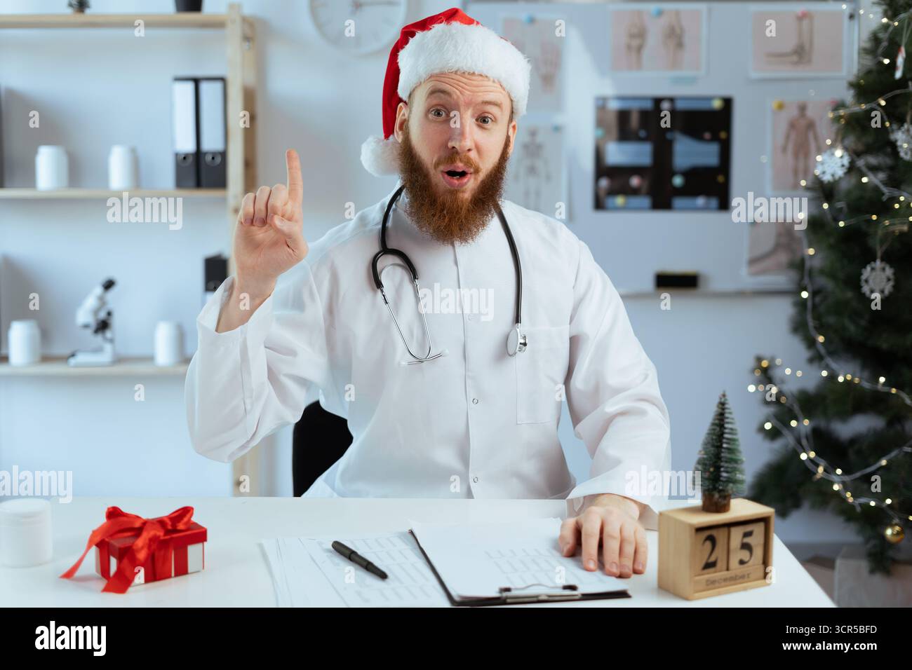 Médecin barbu sur le quart ayant l'idée pointant vers le haut avec le doigt, regardant la caméra tout en travaillant la veille de Noël. copier l'espace Banque D'Images