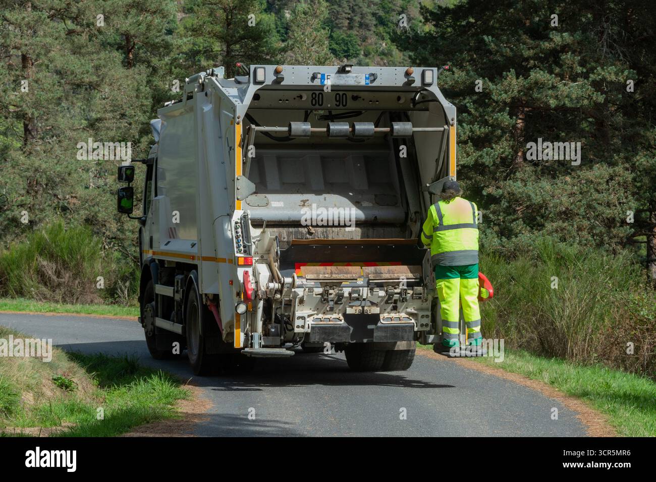 Un camion poubelle roule sur une route rurale sinueuse en Auvergne. Un travailleur portant un uniforme brillant se tient près du camion. France Banque D'Images
